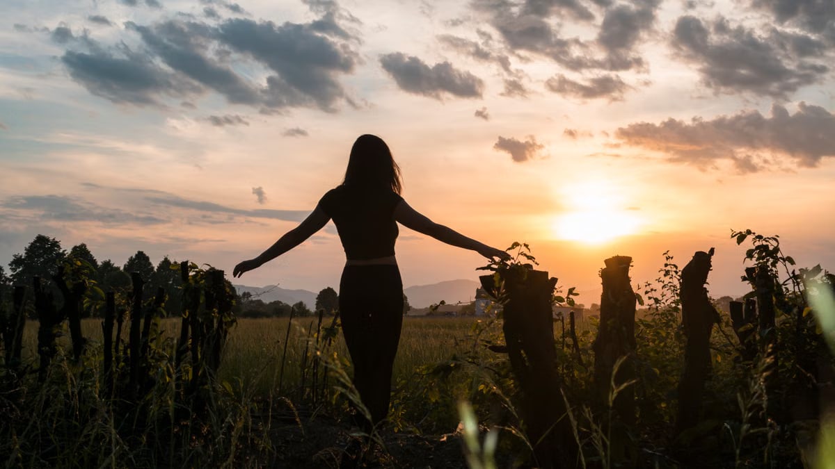 A woman stands in a field at sunset, arms outstretched, capturing the serene moment.